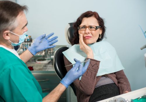 Woman feeling toothache, touching cheek with hand at dental clinic. Senior dentist trying to help. Dental care and health concept