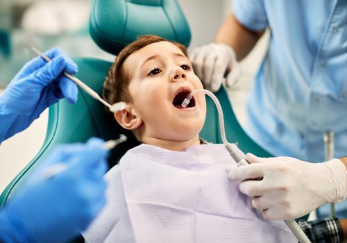 Small boy sitting in dentist's chair with mouth open while getting his teeth checked by dentists at dental clinic.